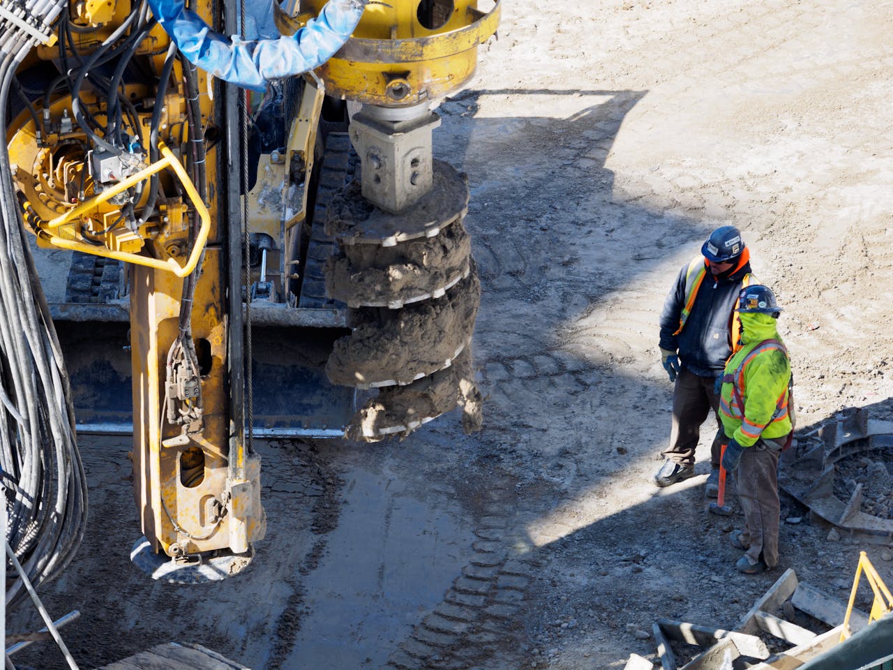 services-03 Two workers in protective gear at a construction site with heavy drilling machinery.