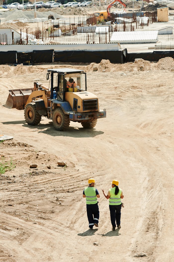 gallery-5 Construction workers in safety gear walking at a busy site with a backhoe in operation.